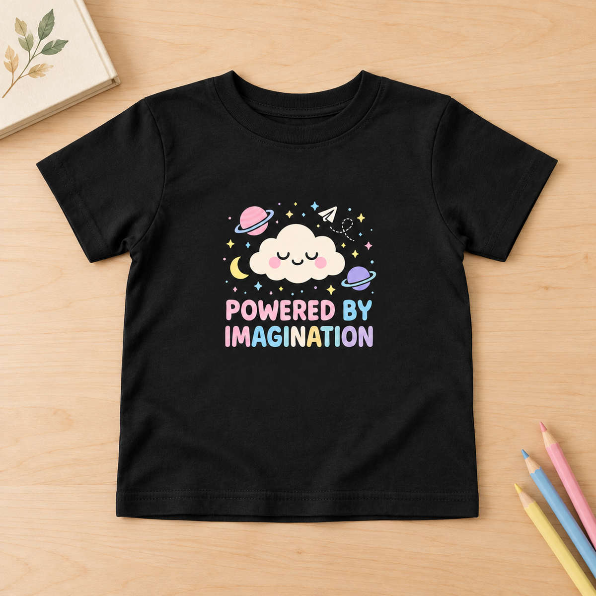 Powered by Imagination black toddler T-shirt, flat lay on light wood with pastel pencils